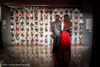 James Corey's Wall of Masks from SEAF 2021. A grey wall with 120 leather masks in various colors hung as an installation at the 2021 Seattle Erotic Art Festival. A couple in black and red stand before it, their backs to the camera.